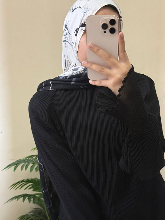 A person taking a mirror selfie wearing a black pleated long-sleeve abaya and a black-and-white patterned hijab, holding a light-colored smartphone.