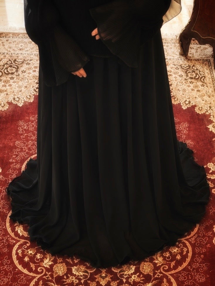 A long flowing black dress or abaya with wide sleeves, shown on a person standing on a red and gold patterned rug indoors.