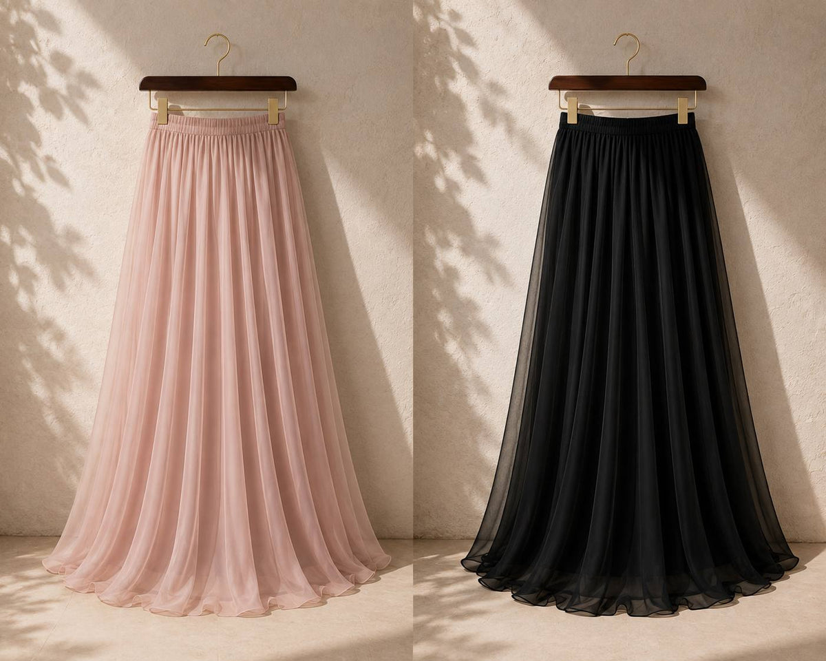 Two long pleated tulle skirts hanging on wooden hangers against a textured beige wall with leaf shadows. The left skirt is light blush pink, and the right skirt is solid black.