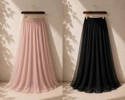 Two long pleated tulle skirts hanging on wooden hangers against a textured beige wall with leaf shadows. The left skirt is light blush pink, and the right skirt is solid black.