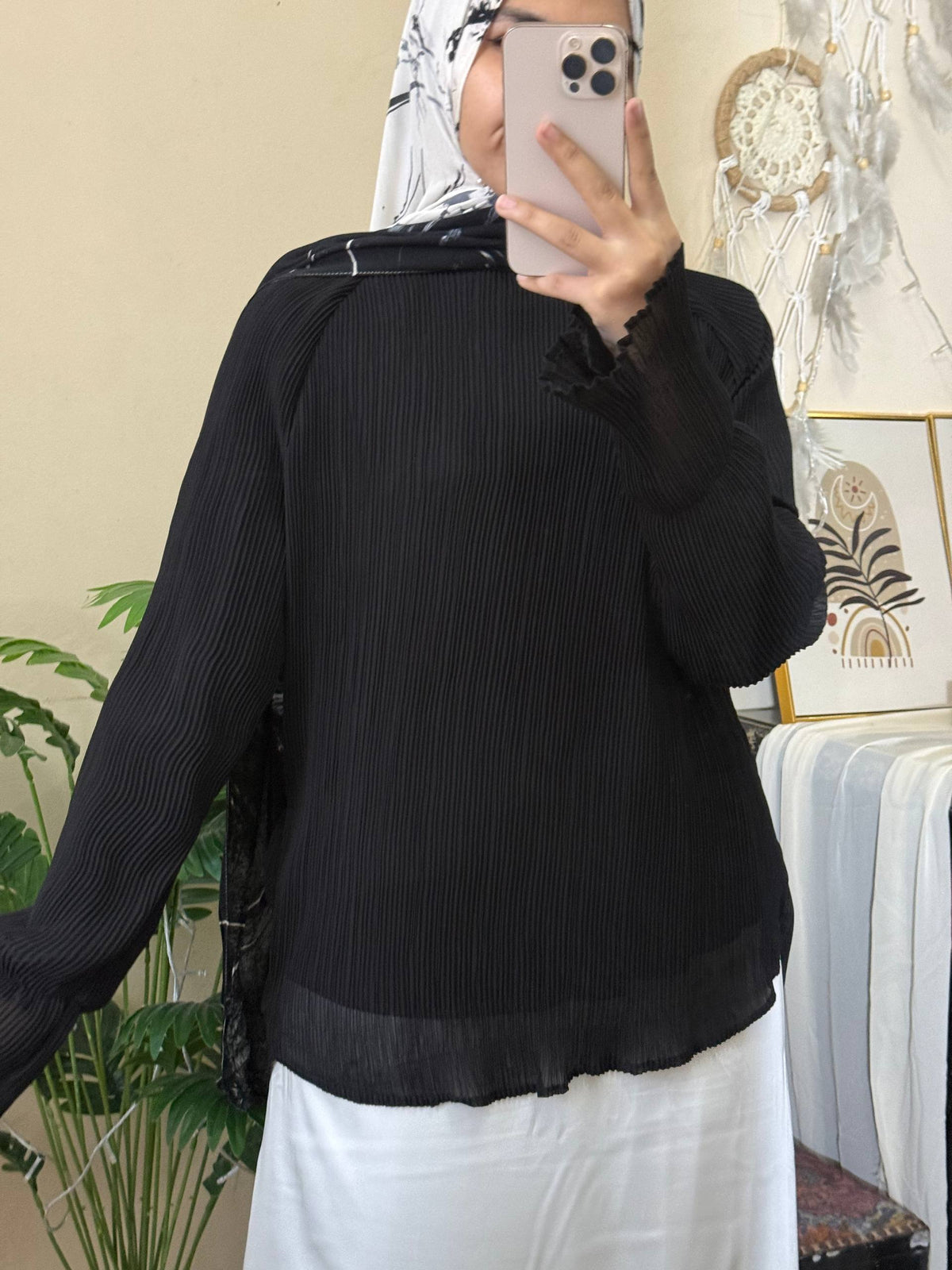 A woman wearing a black long-sleeve pleated blouse with ruffled cuffs and a sheer hem, paired with a white skirt, taking a mirror selfie in an indoor setting.