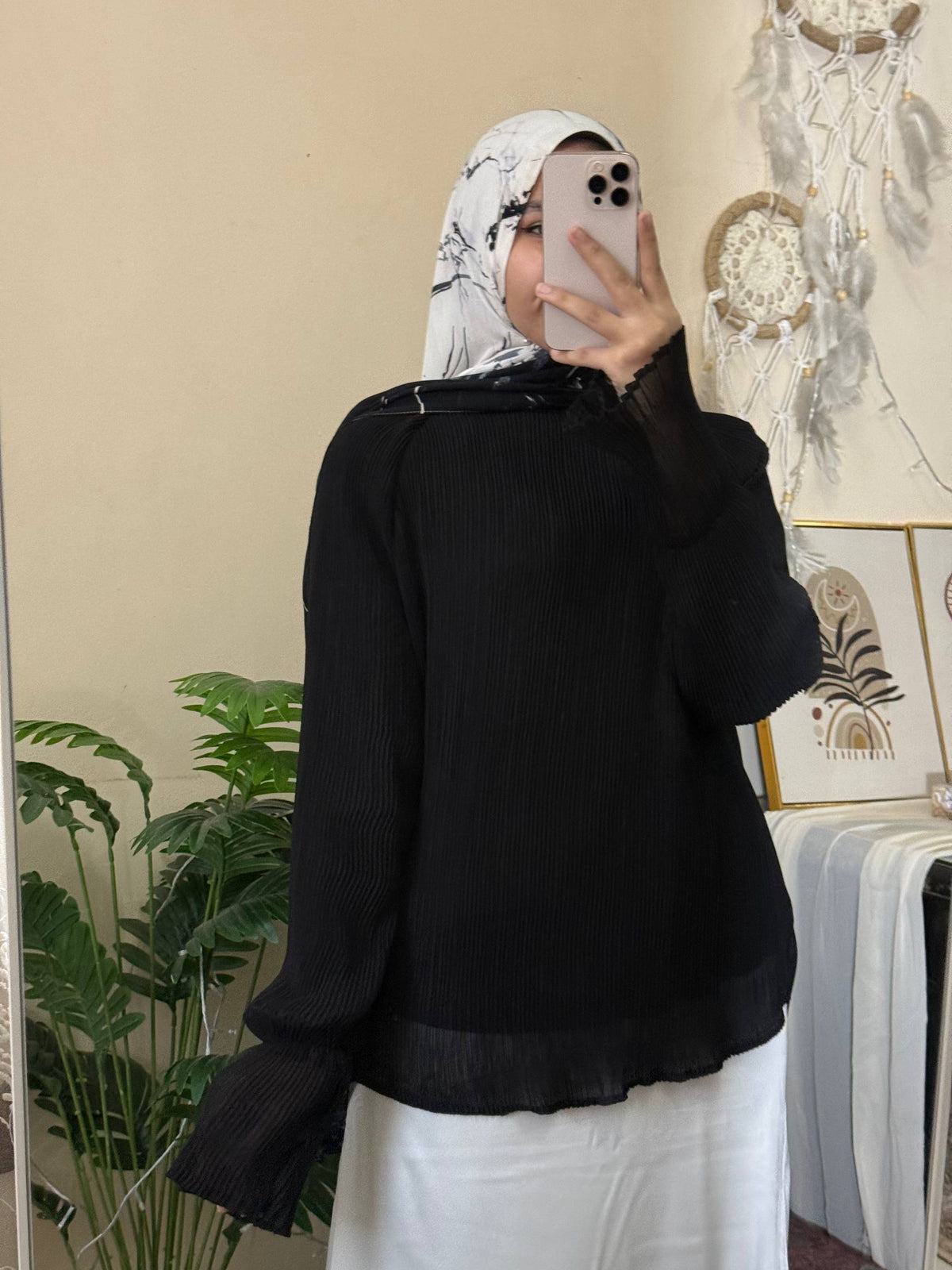 A woman wearing a black long-sleeve ribbed blouse with a sheer ruffled hem and cuffs, paired with a white hijab and white pants, taking a mirror selfie in an indoor setting.