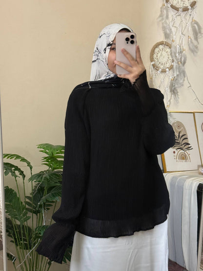 A woman wearing a black long-sleeve ribbed blouse with a sheer ruffled hem and cuffs, paired with a white hijab and white pants, taking a mirror selfie in an indoor setting.