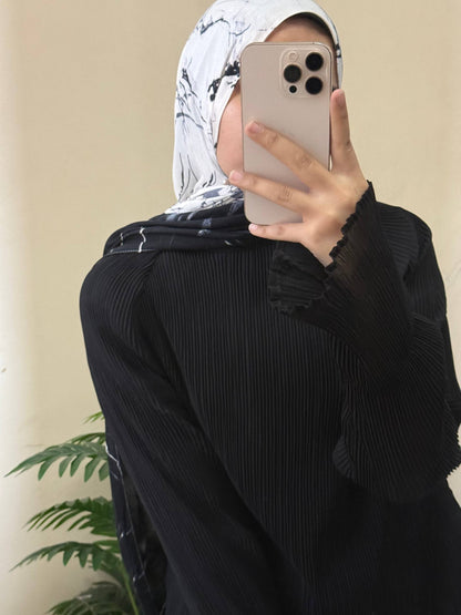 A person taking a mirror selfie wearing a black pleated long-sleeve abaya and a black-and-white patterned hijab, holding a light-colored smartphone.