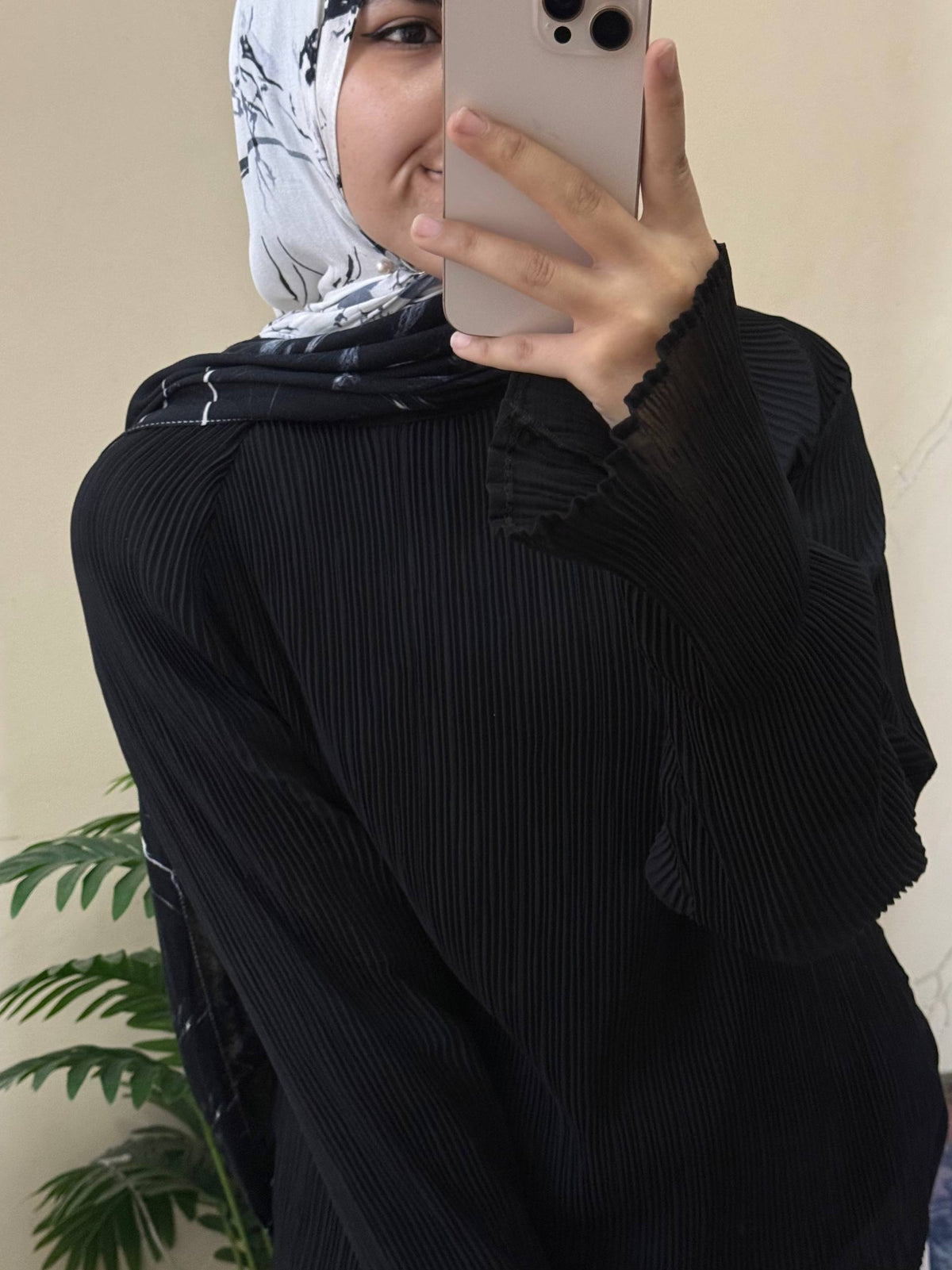 A person taking a mirror selfie wearing a black pleated long-sleeve top with ruffled details on the shoulders and sleeves, paired with a light gray and black patterned hijab.