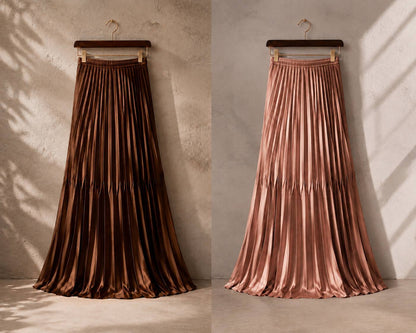Two long pleated satin midi skirts hanging on wooden hangers against a textured concrete wall. The left skirt is dark brown, and the right skirt is a warm metallic rose gold.