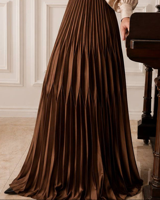 A floor-length pleated brown satin skirt with a train, worn by a model standing in an elegant room next to a dark wooden piano.
