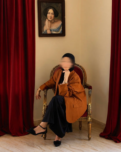 A person seated in an ornate chair between red curtains, wearing a brown blazer, black skirt, and black heels. A framed portrait hangs on the beige wall behind them.