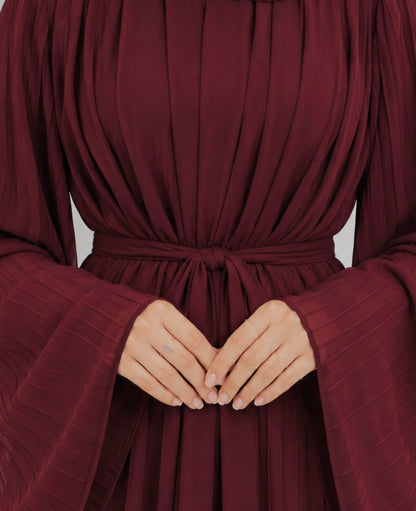 A close-up of a person wearing a deep burgundy long-sleeve dress with pleated fabric, a tie waist belt, and wide flared sleeves. The person's hands are clasped in front, showing a delicate ring on one finger.