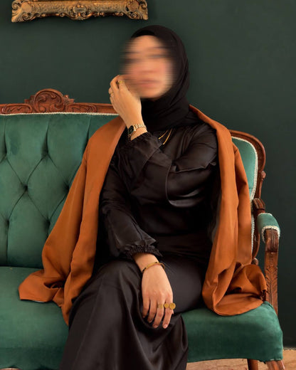 A woman wearing a black abaya and black hijab sits on a green tufted armchair, draped with a rust-orange shawl. The background is a dark green wall with a gold picture frame partially visible at the top.