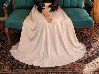 A person seated on a teal upholstered bench wearing a long, flowing champagne satin skirt or gown with a black long-sleeve top. The scene is set on a patterned oriental rug.